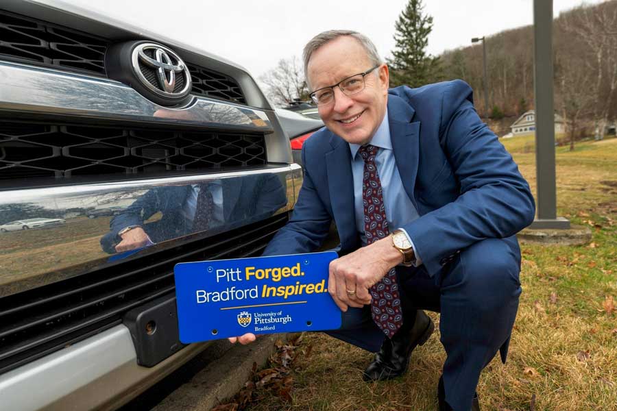 President Rick Esch standing in front of a vehicle holding a license plate that says "Pitt Forged. Bradford Inspired."