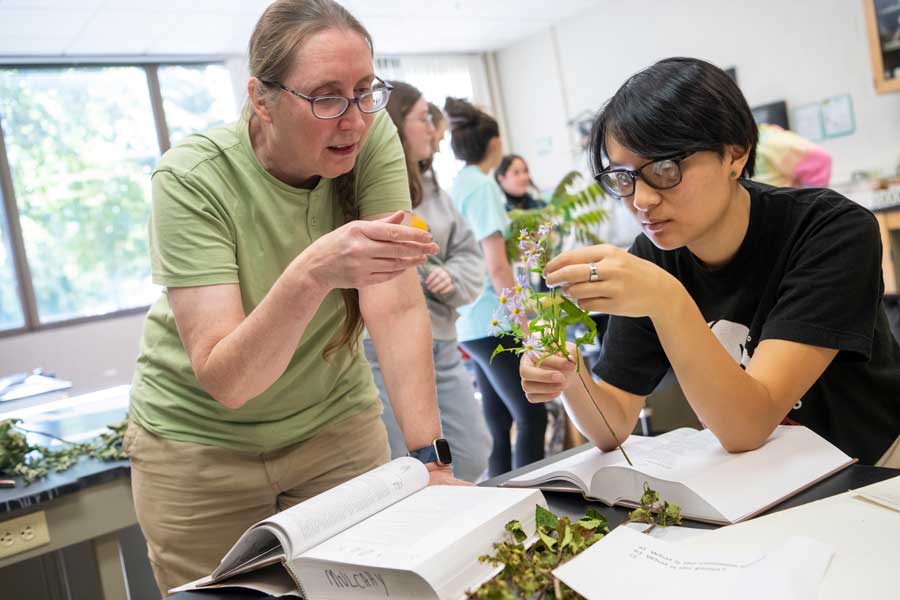 A woman professor leans over a lab table and gestures to a purple-flowered plant that a student is holding. There are several open books on the table to help the two identify the plant.